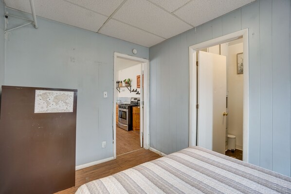 Un dormitorio con cama, piso de madera, puerta que da a una cocina y un reloj en la pared.