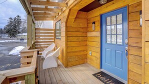 Une cabane en bois avec une porte bleue, une véranda couverte avec des chaises blanches et un paillasson sur lequel est écrit « Bonjour » à l’entrée.