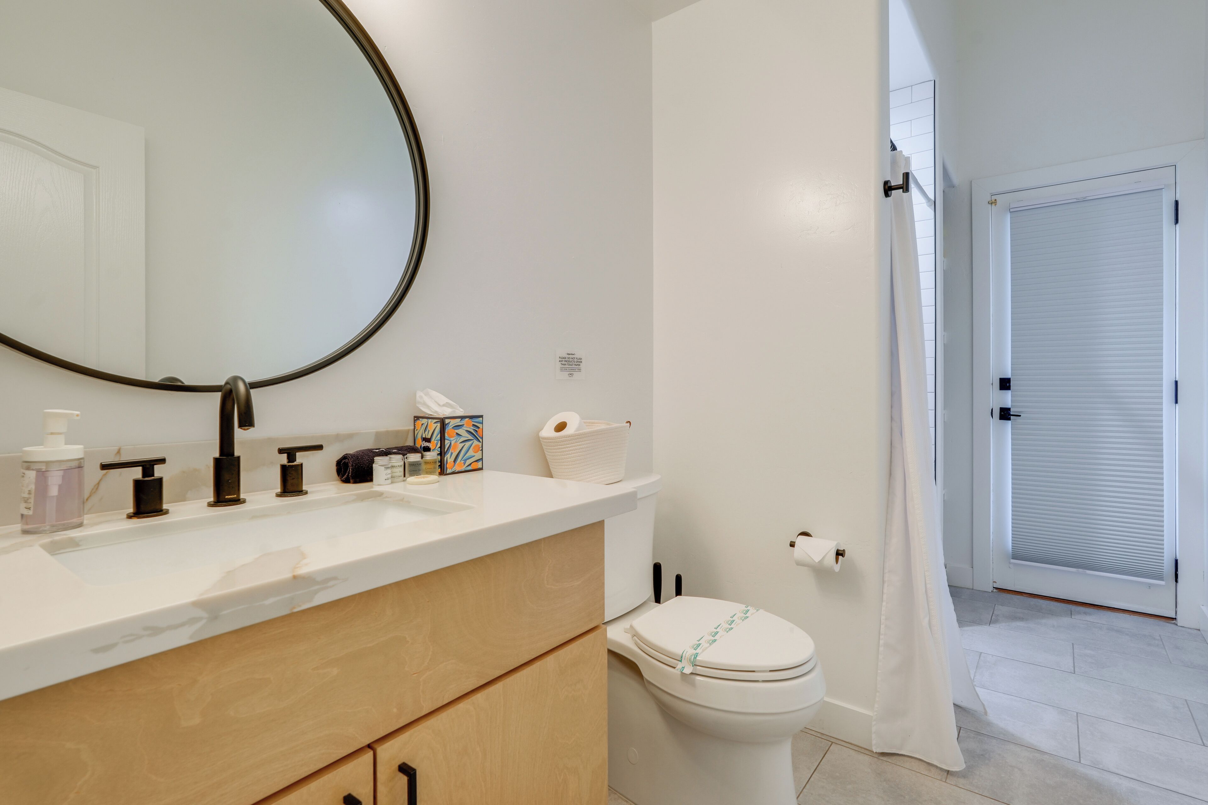 A modern bathroom with a large mirror, white sink, and toilet. There are toiletries on the counter and a towel rack.
