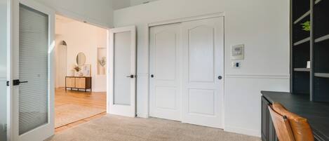 A room with a carpeted floor, white walls, a closed door, a small table with a mirror, and a chair.
