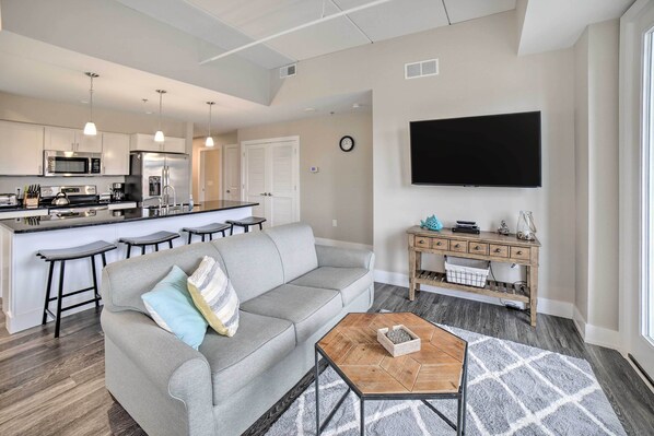 A modern living room with a grey sofa, a coffee table, and a kitchen area with a microwave, refrigerator, and bar stools. - Luxe Manistee Condo w/ Balcony & River Views! (Manistee)