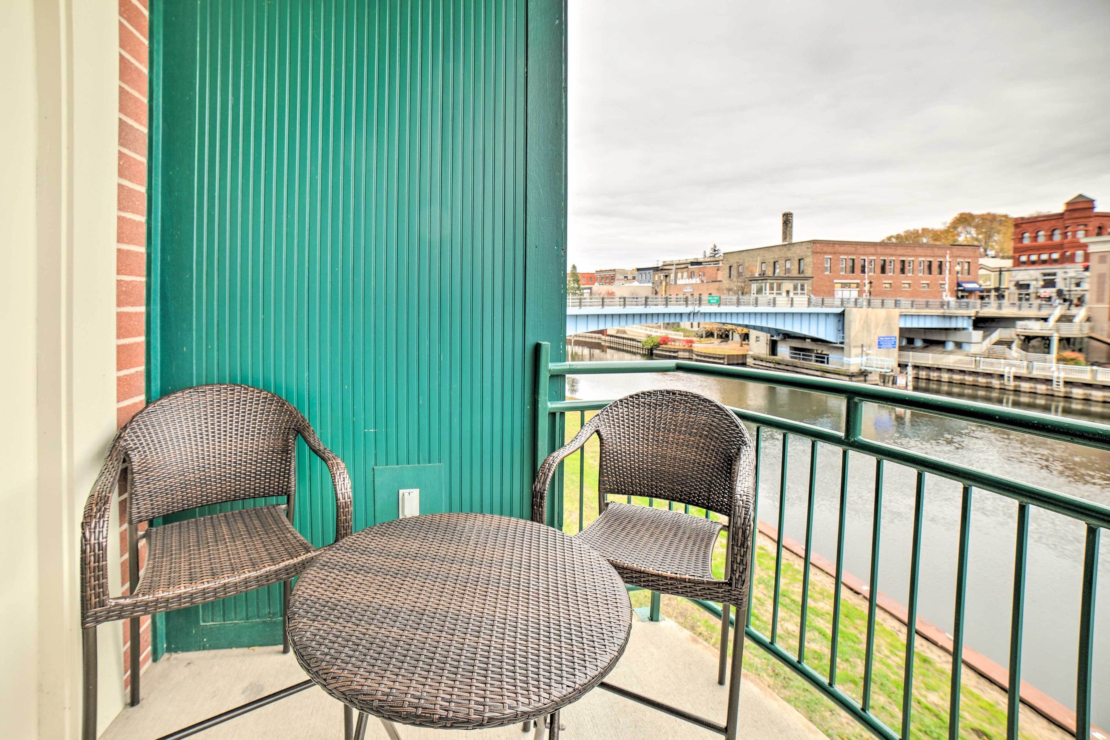 Ein Balkon mit Korbstühlen und einem Tisch, mit Blick auf einen Fluss und Gebäude.