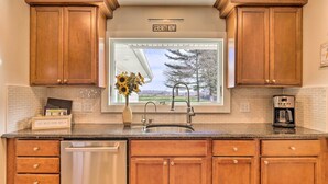 A kitchen with wooden cabinets, a stainless steel sink, and a window with a view of trees and a building. - Mattituck Home w/ Fireplaces - Near Wineries (Mattituck)