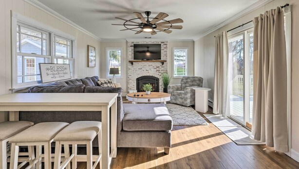 A spacious living room with a fireplace, a ceiling fan, and a large window with curtains. - Mattituck Home w/ Fireplaces - Near Wineries (Mattituck)