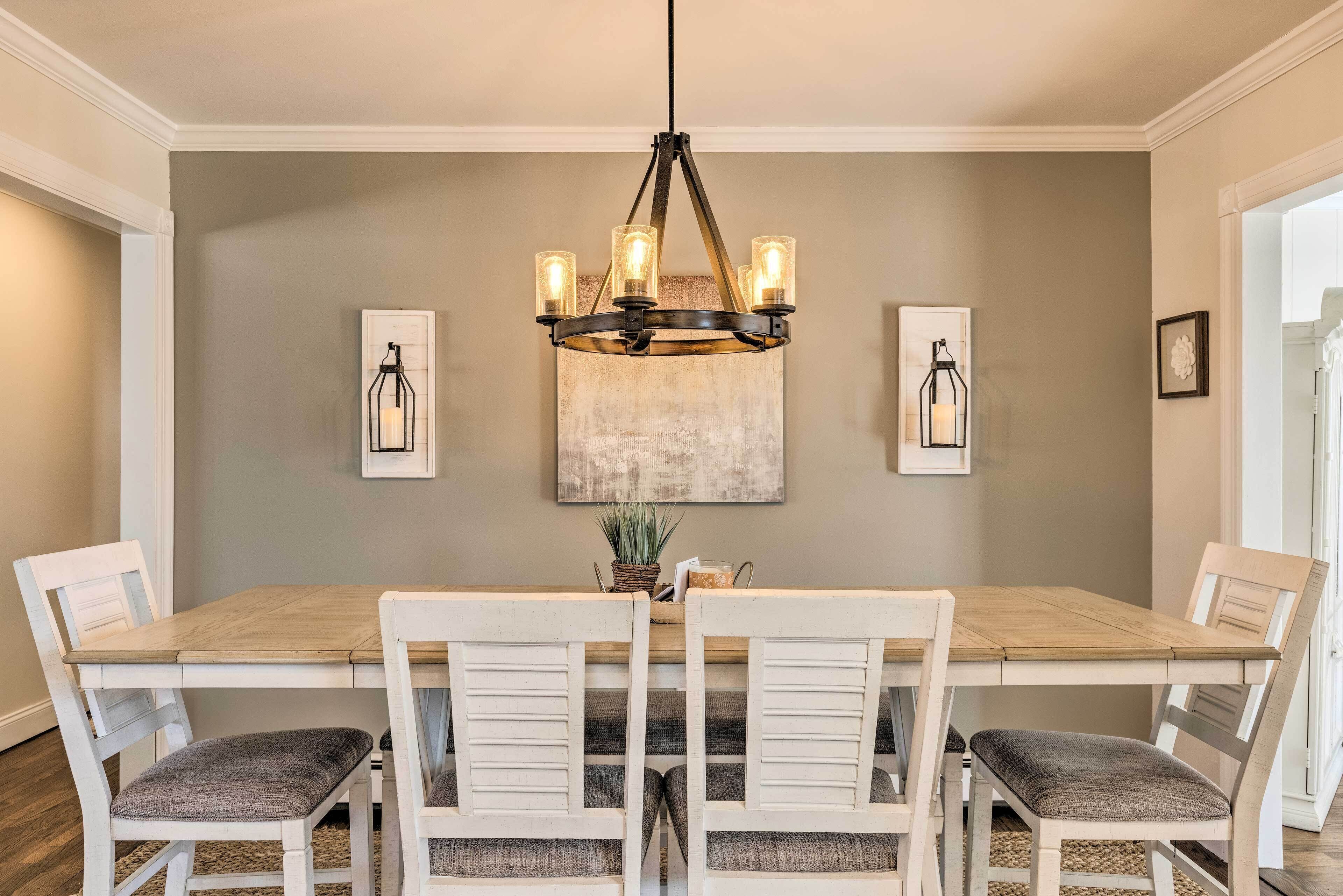 Une salle à manger avec une table en bois, des chaises blanches, un lustre et des œuvres d’art encadrées accrochées au mur.