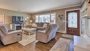 A living room with a gray sofa set, a coffee table, and a TV. There is a wooden floor, a rug, and a door with a window. - Mattituck Home w/ Fireplaces - Near Wineries (Mattituck)