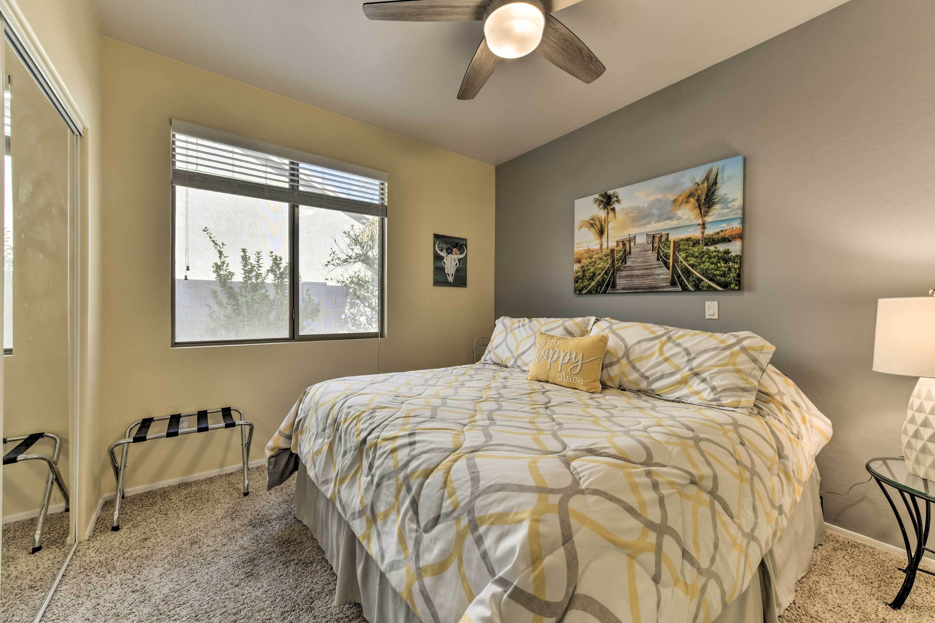 A bedroom with a bed, a ceiling fan, a window, a painting, and a bench.
