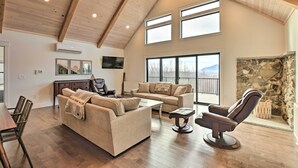 Amplia sala de estar con techo alto, vigas de madera, chimenea de piedra y grandes ventanales con vistas al exterior.