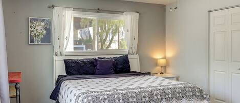 A bedroom with a bed, a window with curtains, a ceiling fan, a bedside lamp, and a door.