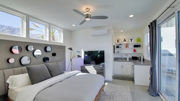 A modern bedroom with a bed, a ceiling fan, a TV, a kitchenette, and a large window. - Modern Oasis w/ Mtn-view Pool Deck - Walk Downtown (Palm Springs)