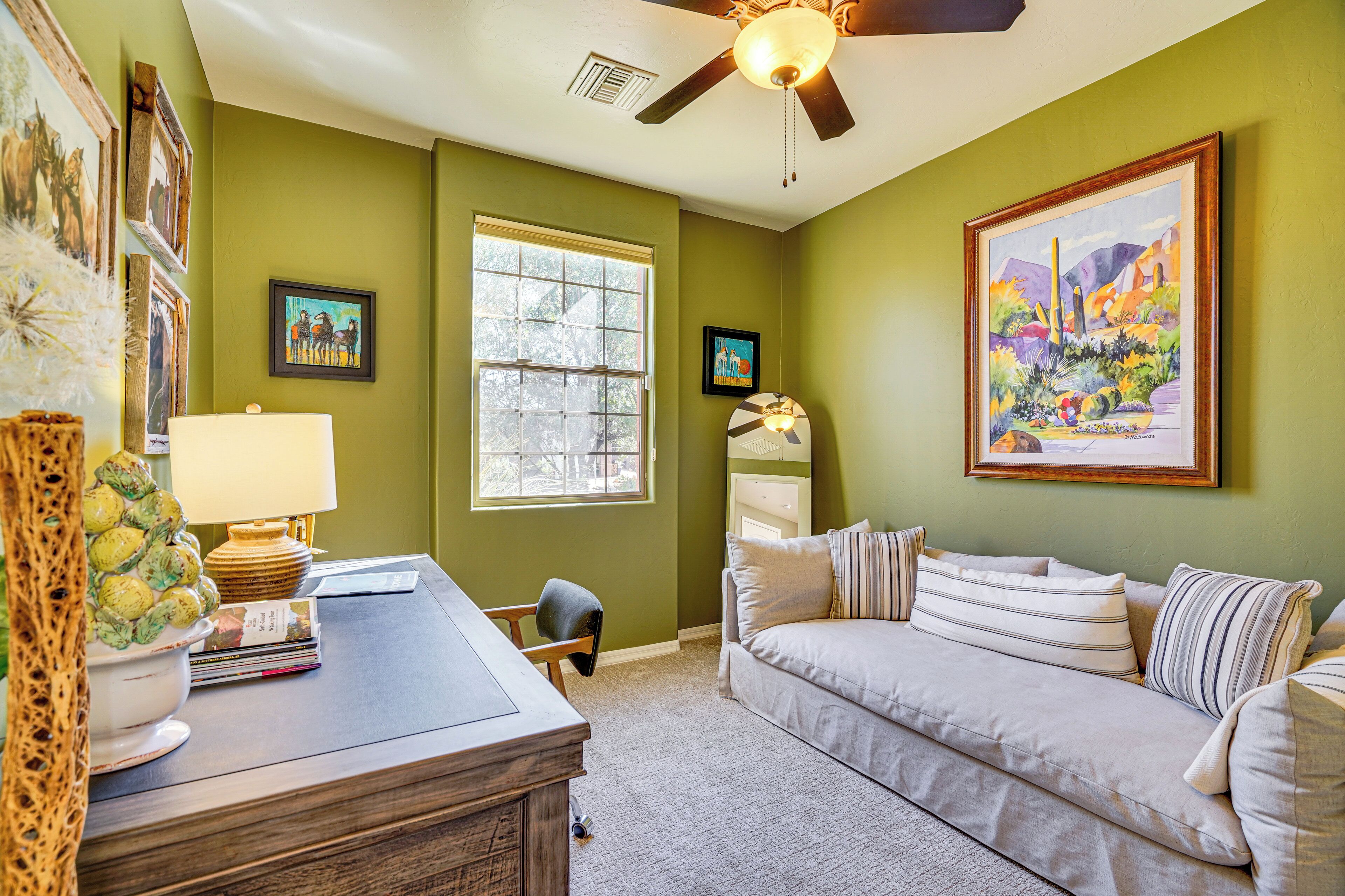 A room with a green wall, a ceiling fan, a window, a couch, and a desk with a lamp and books.