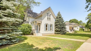 A two-story house with a front porch, surrounded by well-maintained lawns and evergreen trees. - Walk to Lake Michigan & Dtwn: Coastal Grand' (Ludington)