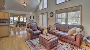 A spacious living room with a brown leather sofa, a wooden coffee table, and a large window with a view of trees. - Deck, Pool, Trails Access: Pocono Family Home! (Albrightsville)