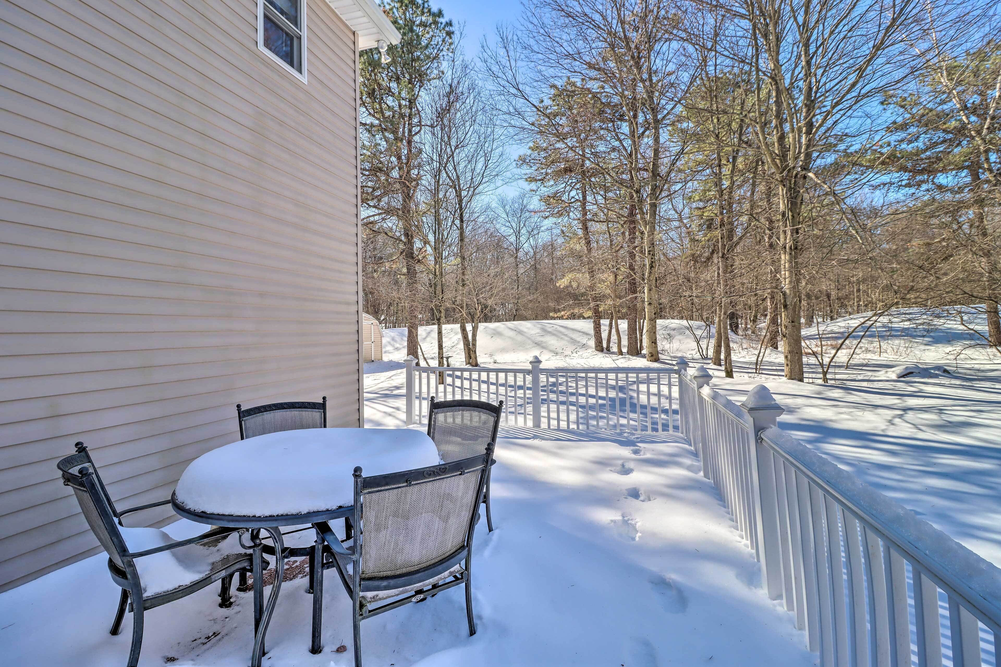A snow-covered patio with a table and chairs, a white fence, and leafless trees in the background.