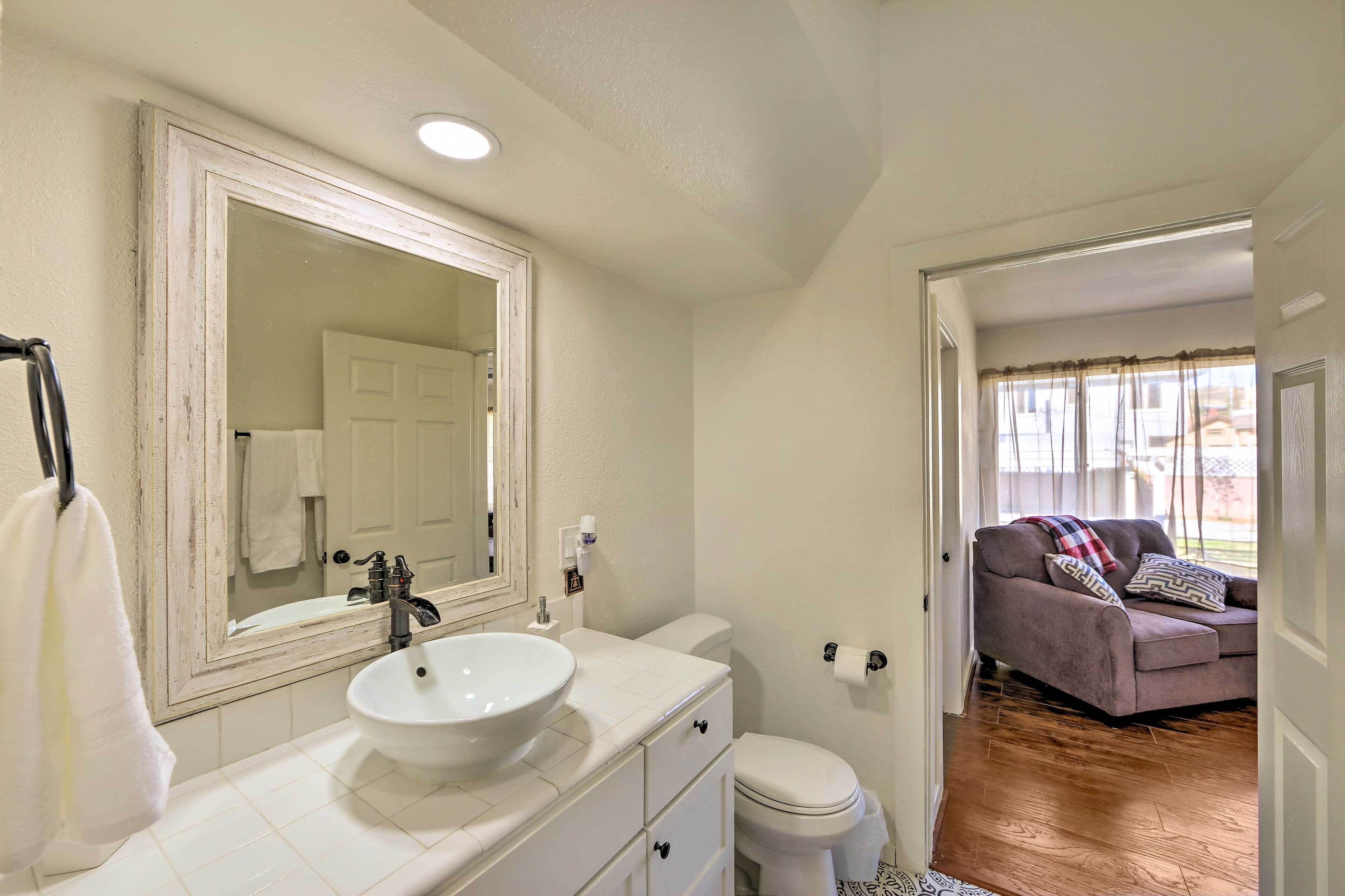 A bathroom with a white sink, mirror, and toilet. There is a couch in the adjacent room.