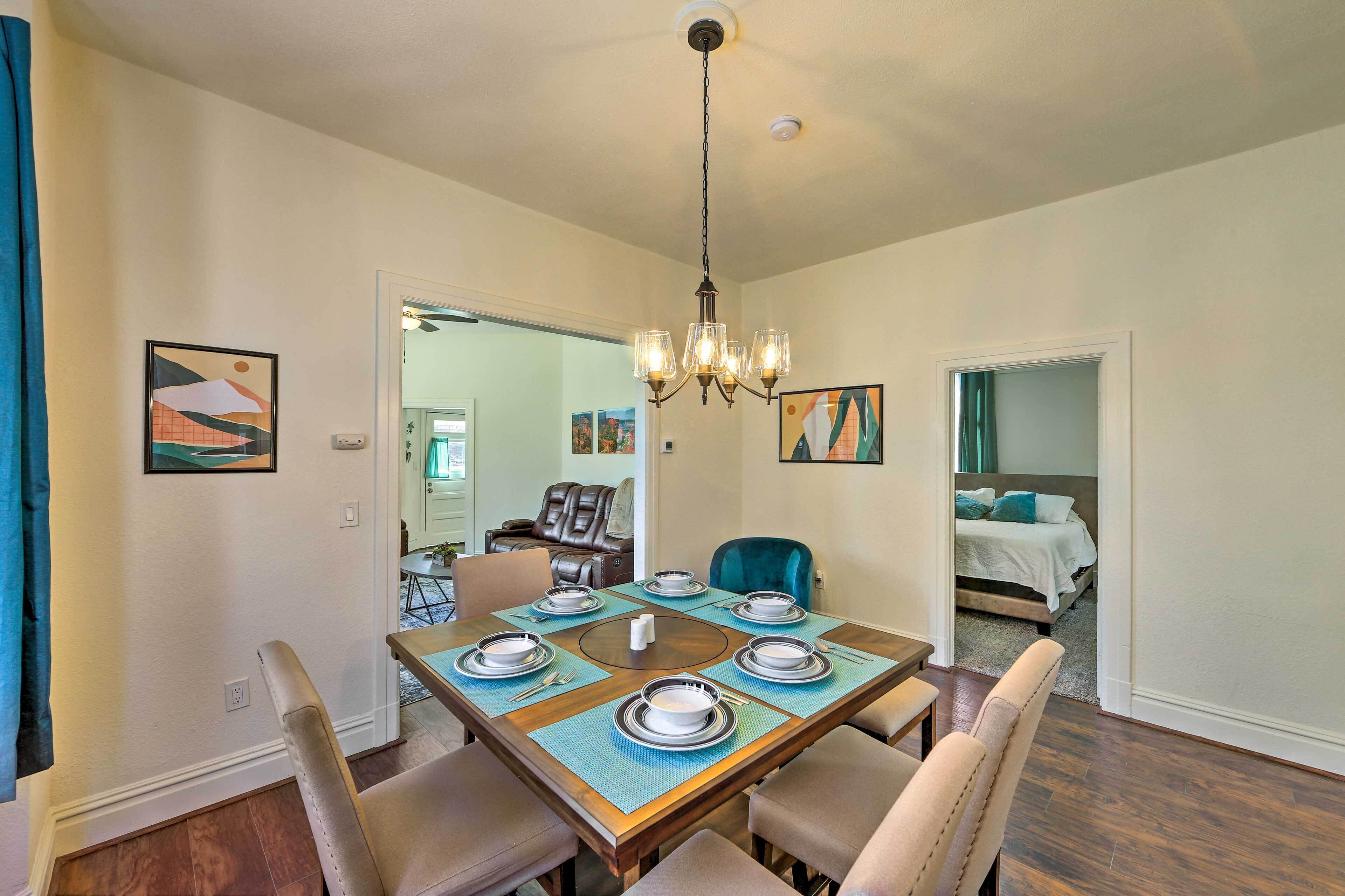 A dining area with a wooden table set for six, surrounded by six chairs. A chandelier hangs from the ceiling, and there are two paintings on the walls.
