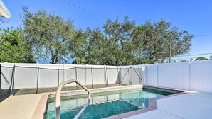 Une piscine rectangulaire avec un escalier en métal, entourée d’une clôture blanche et bordée d’arbres en arrière-plan.