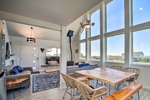 A spacious living area with a large wooden dining table, wicker chairs, a grey sofa, and a blue rug. - Deck + Walk to Beach: Southern Oregon Coast Home! (Gold Beach)