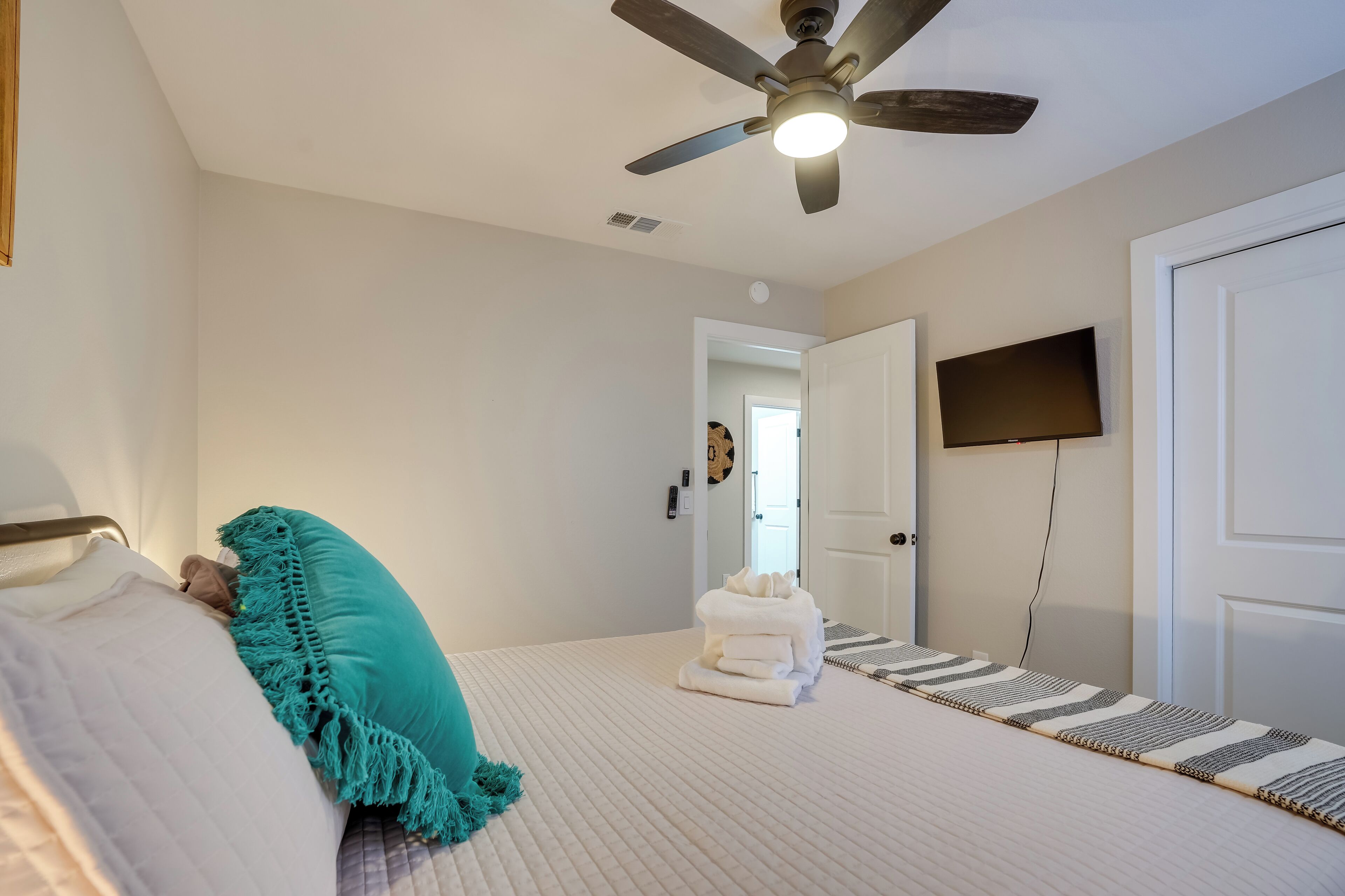 A bedroom with a bed, a ceiling fan, a TV mounted on the wall, and a door leading to another room.