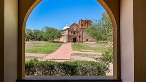 A view through an arched window of a historic building with a central dome and a bell tower, surrounded by a well-maintained lawn and trees. - Adobe-style Tumacacori Home w/ Trail Access! (Tumacacori)