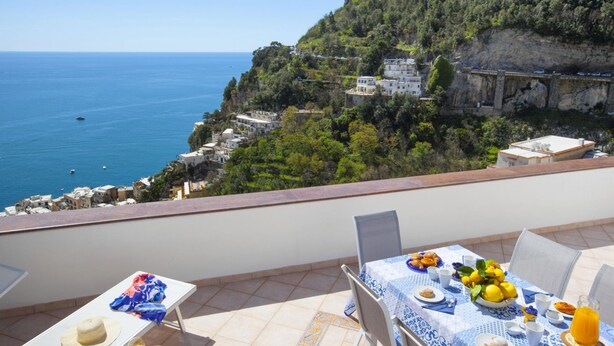 Egy erkély, ahol egy asztal van megterítve a reggelihez, és ahonnan a tengerpartra, valamint a sziklákon épült házakra nyílik kilátás.