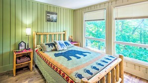 Ein Zimmer mit einem Bett, ausgestattet mit einer Bettdecke mit Tiermotiven, einem hölzernen Kopfteil, einem Nachttisch mit Lampe und einem Fenster mit Blick auf Bäume.