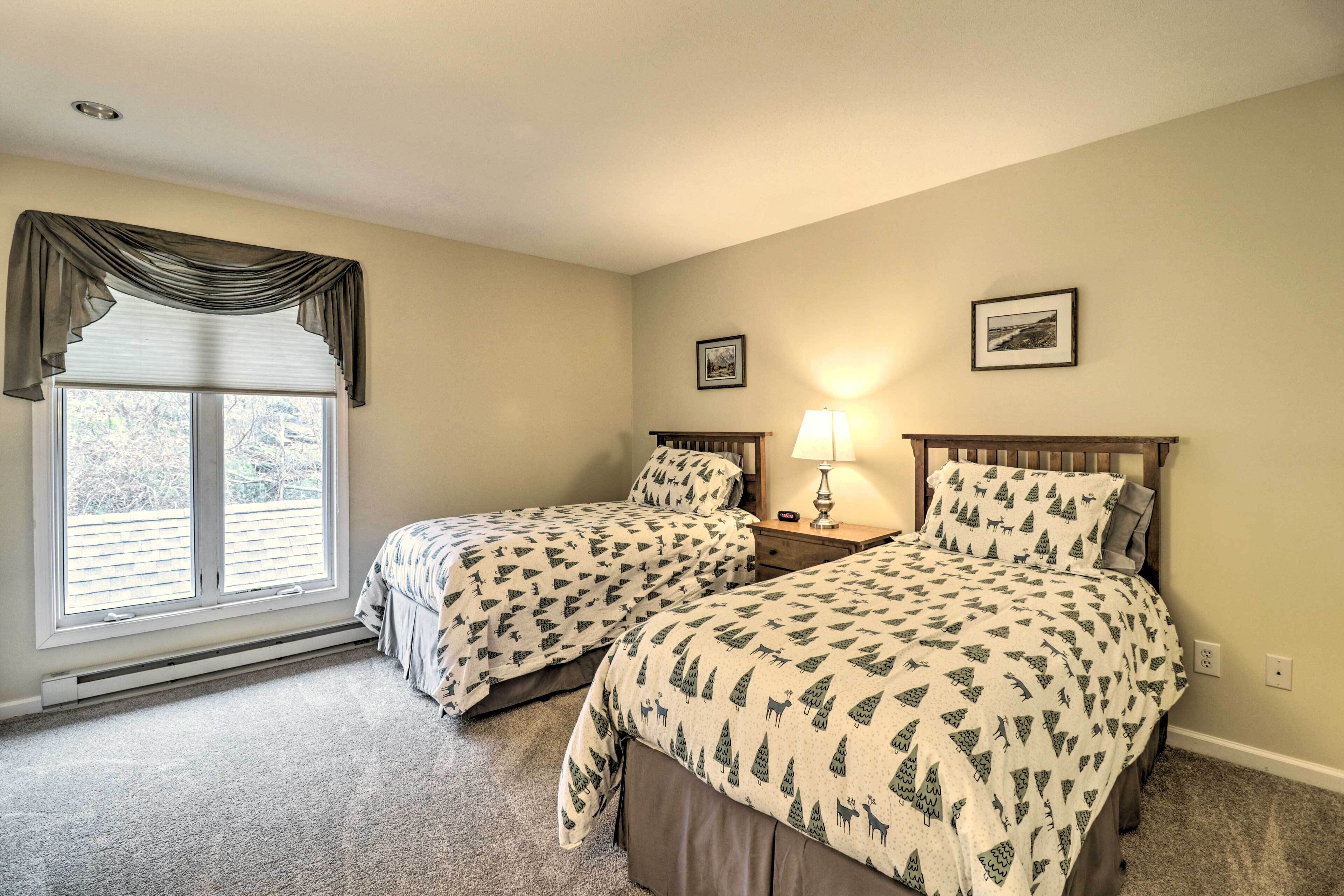 A bedroom with two beds, a window with blinds, a lamp on a nightstand, and two framed pictures on the wall.