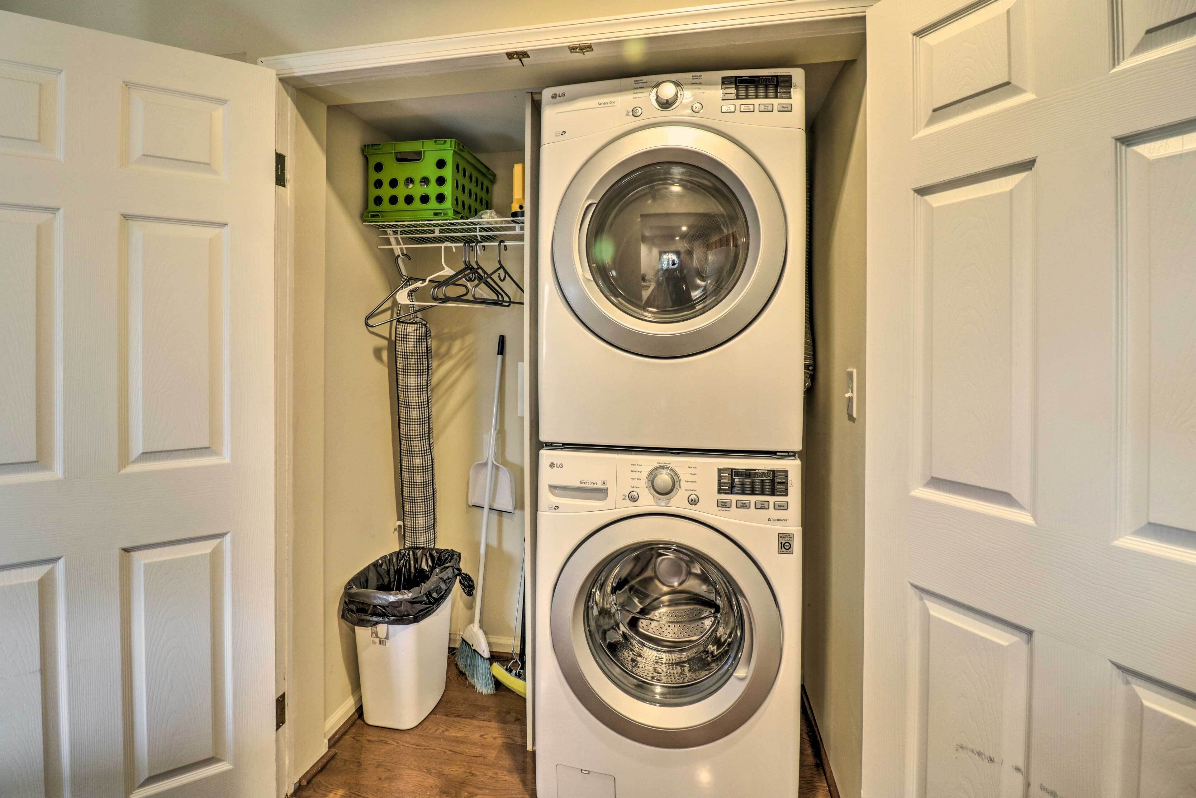 A laundry room with two stacked washing machines, a green basket on a shelf, a broom, and a trash bin.