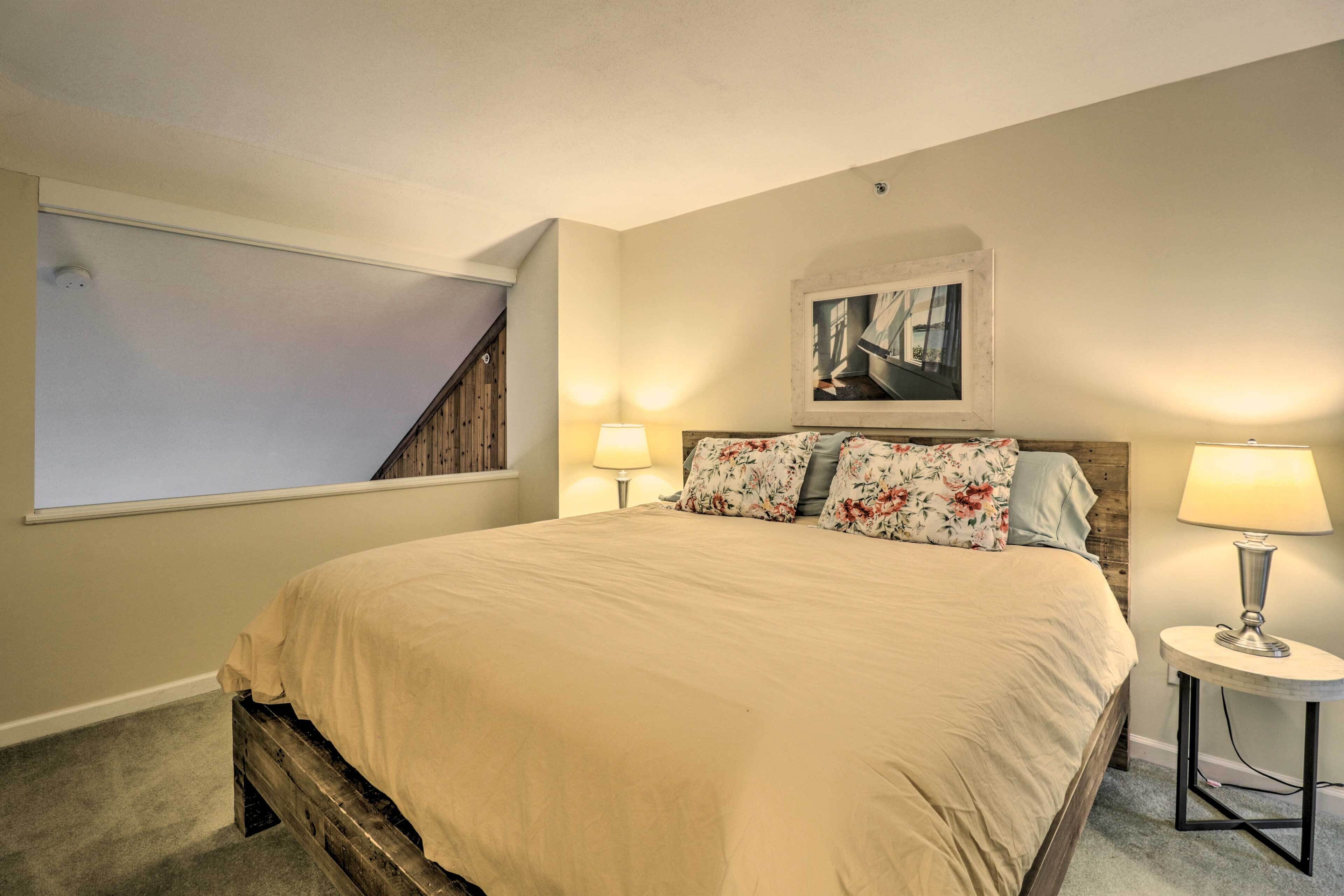 A bedroom with a large bed, two bedside lamps, a framed picture above the bed, and a window with a view of a staircase.