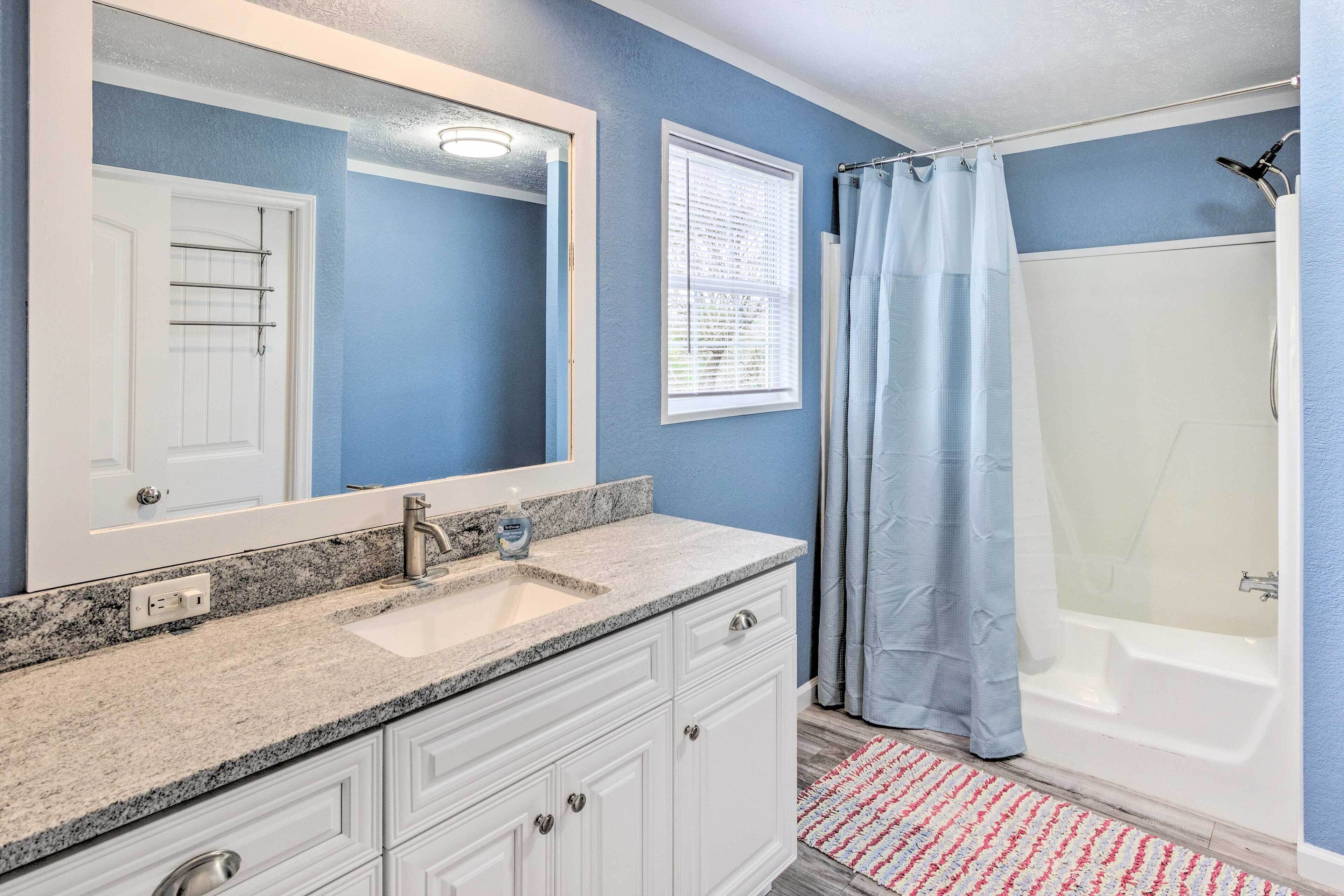 A bathroom with a granite countertop sink, white cabinets, a large mirror, a shower with a curtain, and a small rug on the floor.