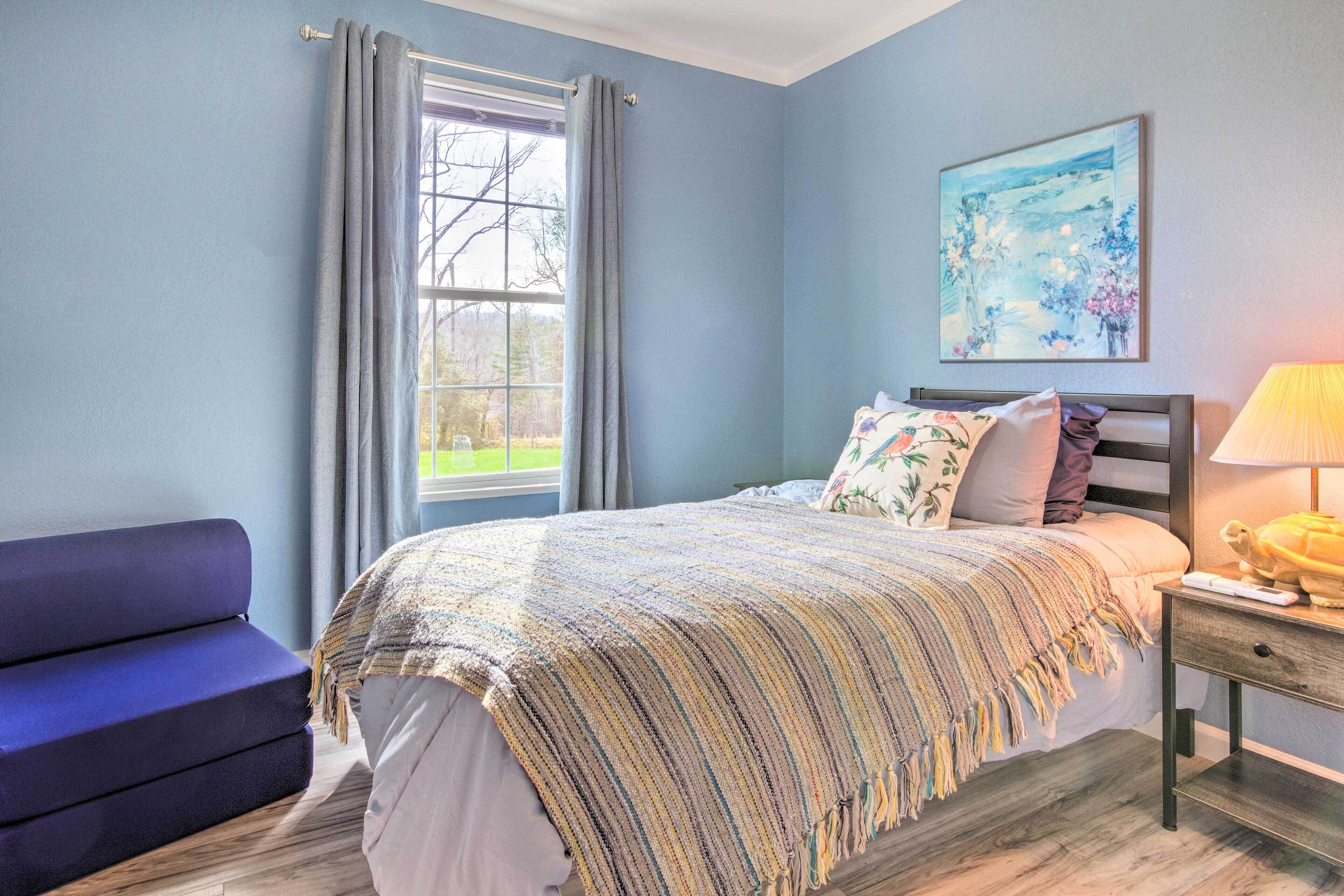 Een slaapkamer met een bed, een nachtkastje, een lamp, een raam met gordijnen, een bank en een schilderij aan de muur.