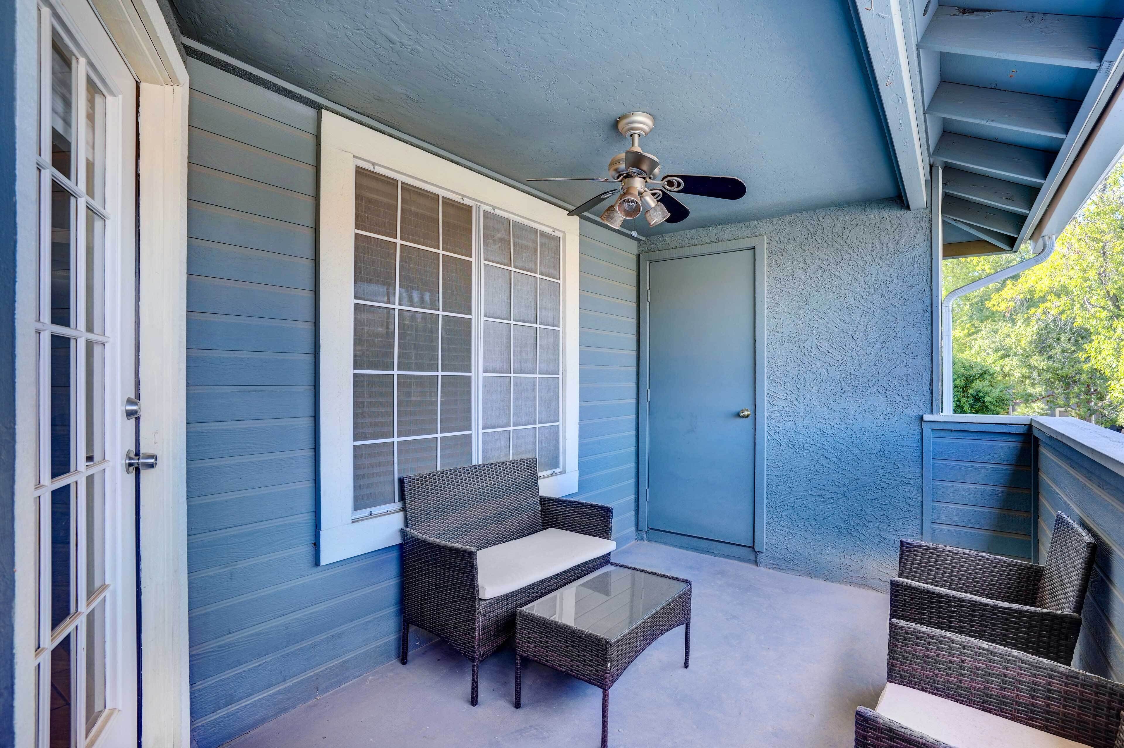 Une véranda couverte avec un ventilateur de plafond, des meubles en osier et une table en verre.