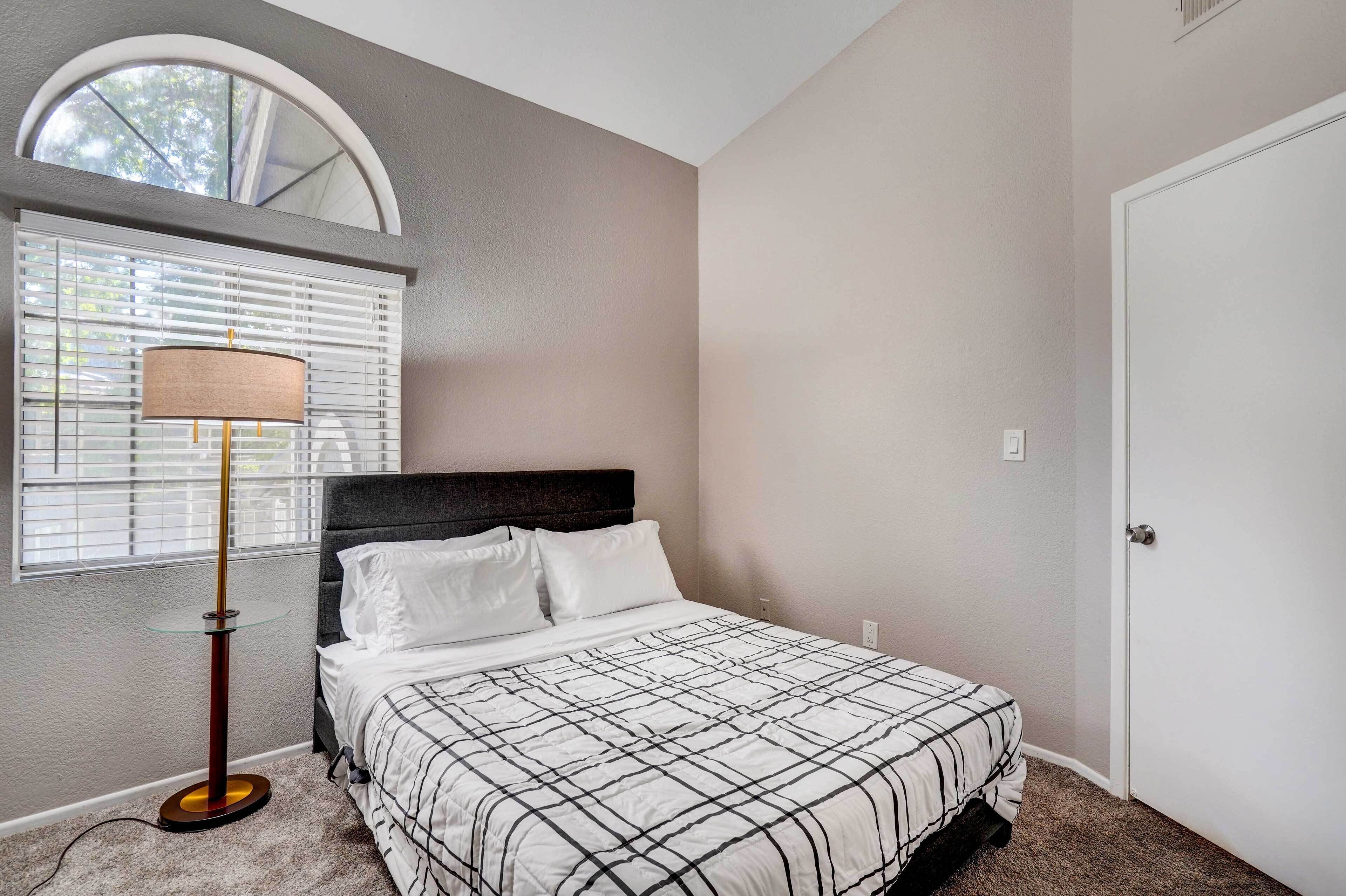 A bedroom with a bed, a floor lamp, a window with blinds, and a door.