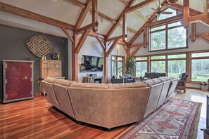 A spacious living room with a large L-shaped sectional sofa, wooden beams on the ceiling, and large windows offering a view of the outdoors.