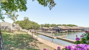A waterfront area with a dock, a building with a flagpole, and a grassy area with purple flowers.