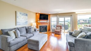 A spacious living room with a fireplace, grey sofas, and a large window offering a view of the outdoors.