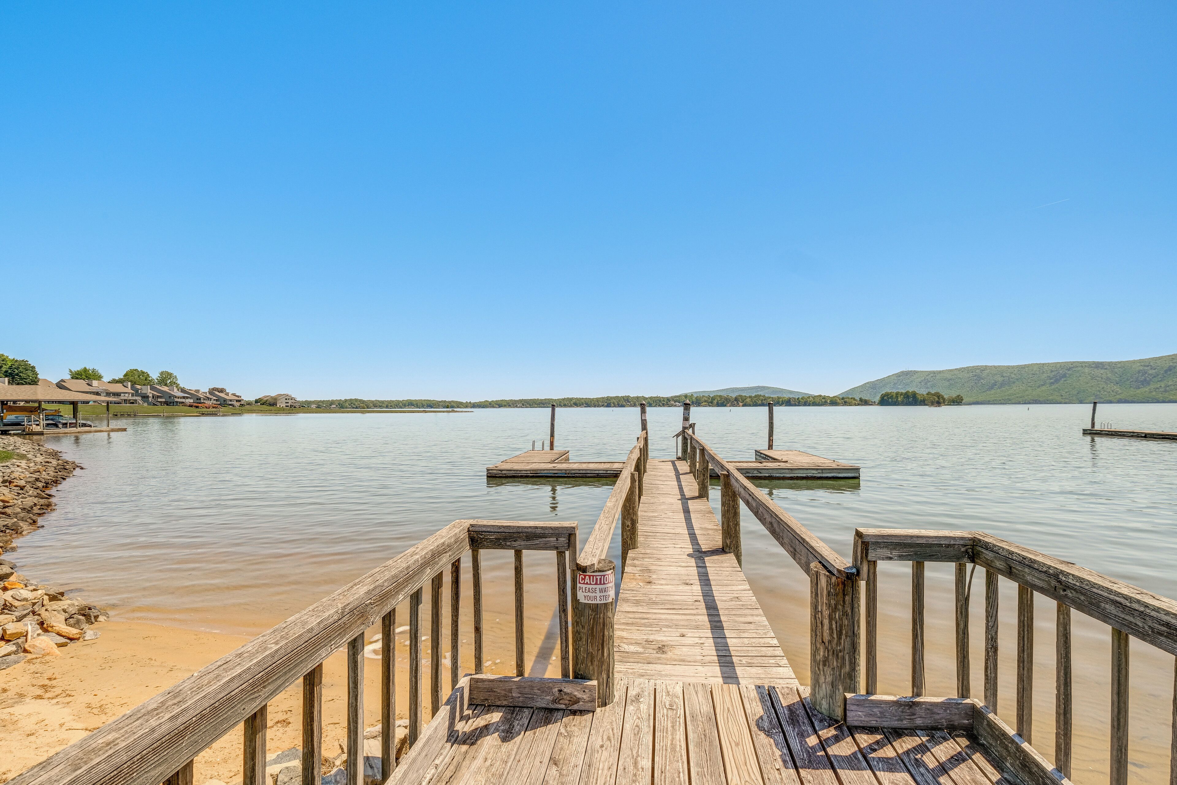 A wooden pier extending into calm water with a 'CAUTION' sign, surrounded by a clear sky and distant hills.