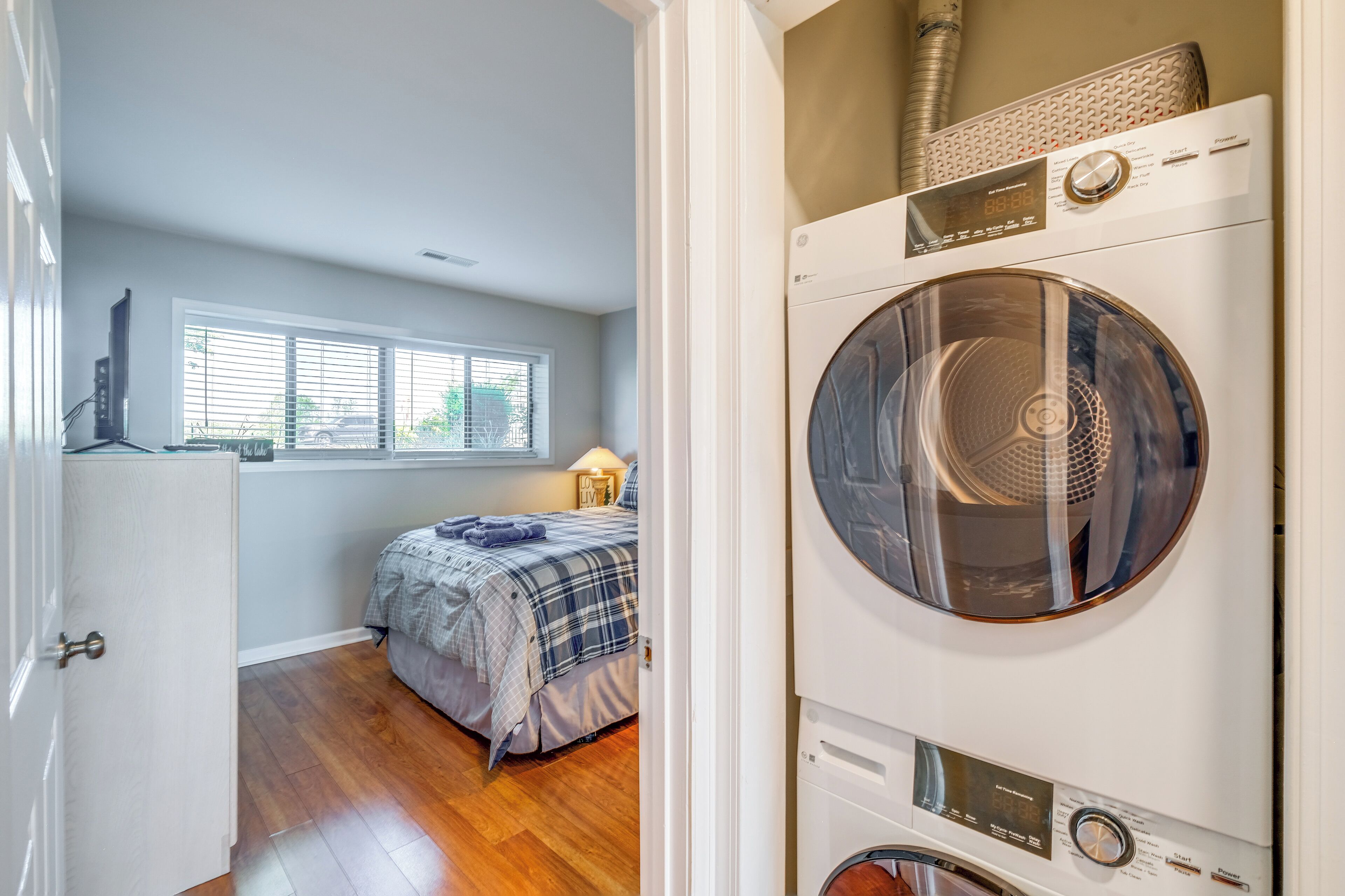 Une buanderie équipée d’un lave-linge et d’un sèche-linge à chargement frontal, un lit avec une couette à carreaux et une fenêtre avec des stores.