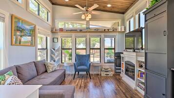 A spacious living room with a gray sofa, a blue armchair, and a fireplace. There are large windows and a ceiling fan.