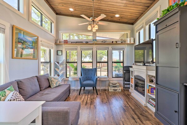 A spacious living room with a gray sofa, a blue armchair, and a fireplace. There are large windows and a ceiling fan. - Upscale Tiny Home - Boho-chic Austin Getaway! (Austin)