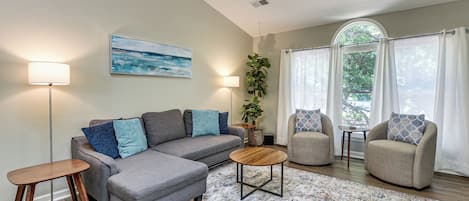 A living room with a grey sectional sofa, two armchairs, a coffee table, a floor lamp, a side table, a potted plant, and a painting on the wall.