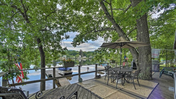 A wooden deck with outdoor furniture, a flag, and a view of a lake and buildings in the distance. - Riverfront Escape w/ Dock: 19 Mi to Saratoga! (Lake Luzerne)