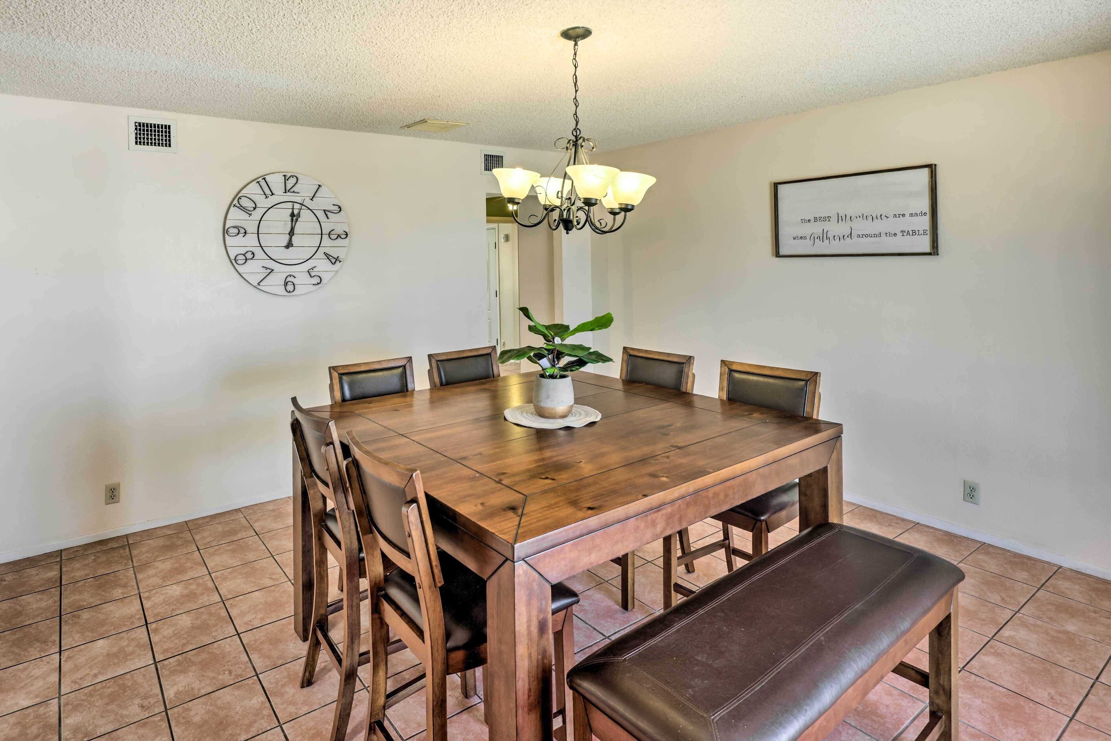 A dining room with a wooden table, chairs, a bench, a clock, a chandelier, and a framed quote on the wall.