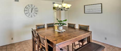 A dining room with a wooden table, chairs, a bench, a clock, a chandelier, and a framed quote on the wall.