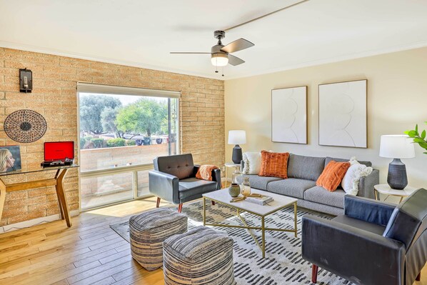 A modern living room with a brick wall, a ceiling fan, a glass desk with a red object, a grey sofa with orange pillows, and a patterned rug.