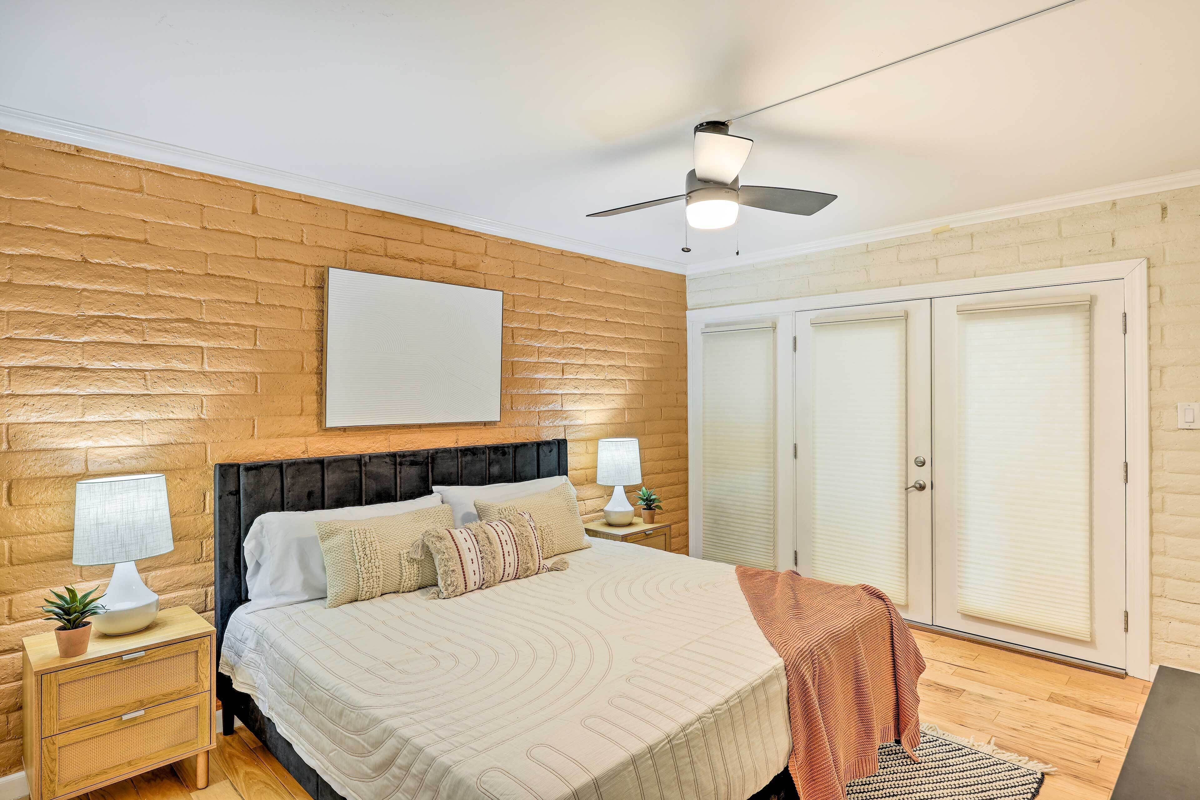A bedroom with a bed, two bedside tables with lamps, a ceiling fan, a large wardrobe, and a brick wall.