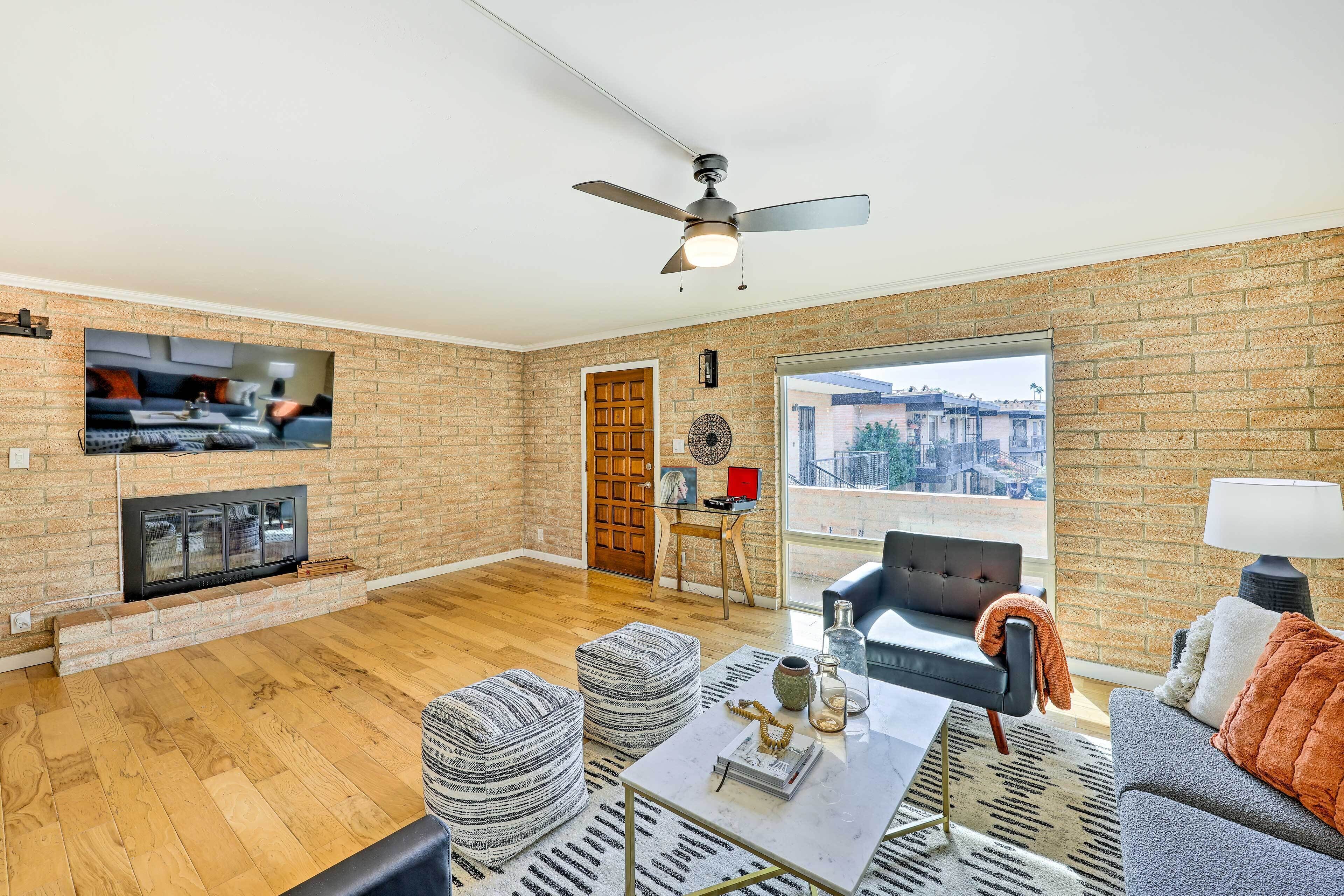 A spacious living room with a fireplace, a ceiling fan, and a large window offering a view of buildings.