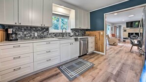 A modern kitchen with white cabinets, a stone backsplash, and a dishwasher. There is a window providing natural light, and a dining area with a TV and grill in the background. - Secluded Suttons Bay Retreat w/ Fire Pit! (Suttons Bay)