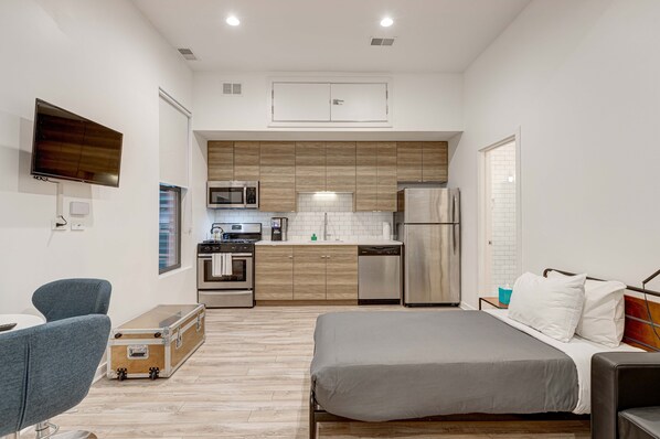 Quarto de hotel compacto com cama, pequeno espaço de cozinha, televisão e mala.