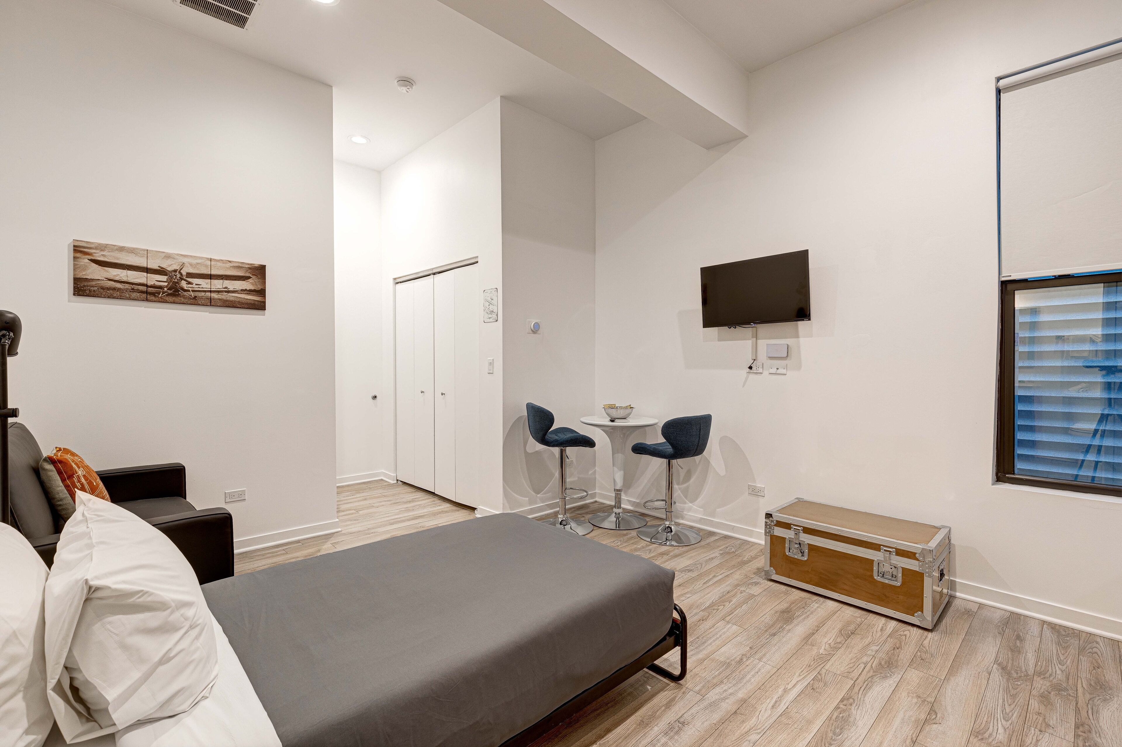 A modern hotel room with a bed, a desk, two chairs, a TV, and a window with blinds.